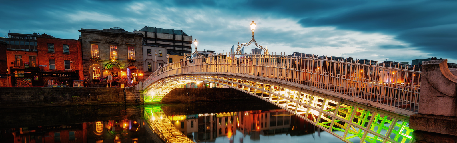 A bridge with green and white lights leads to several brick buildings on a cloudy night.